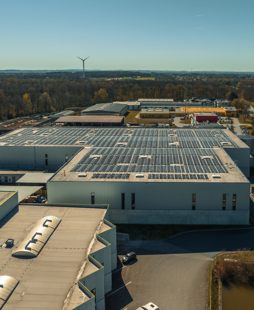 Industriegebäude mit Solarpaneelen bedeckt, umgeben von Bäumen, Straßen und einem Windrad in der Ferne.