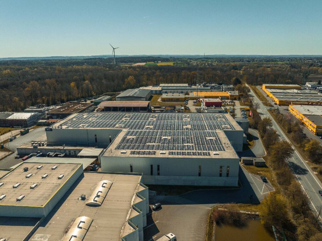 Dach des Industriegebäudes, bedeckt mit Solarpaneelen, in landschaftlicher Umgebung mit Windturbine.