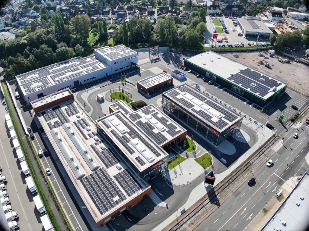 Gebäude mit Solarpanelen steht auf einem Parkplatz, umgeben von Straßen und grünen Flächen.