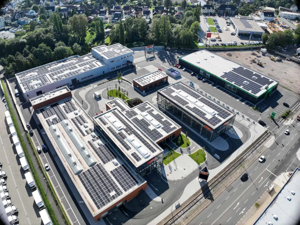 Gebäude mit Solarpanelen steht auf einem Parkplatz, umgeben von Straßen und grünen Flächen.