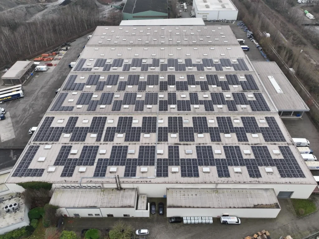 Dach mit Solarpanelen bedeckt, auf großem Industriegebäude, umgeben von Bäumen und Fahrzeugen.