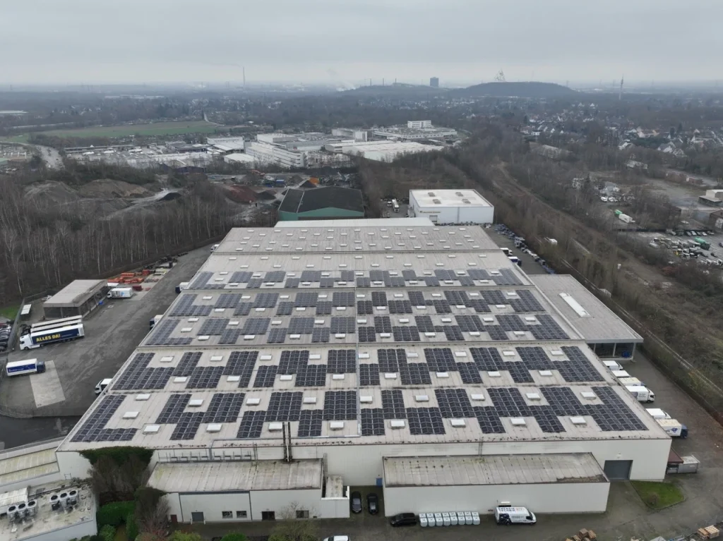 Dach mit Solarpanelen bedeckt, auf großem Industriegebäude, umgeben von Bäumen und Fahrzeugen.