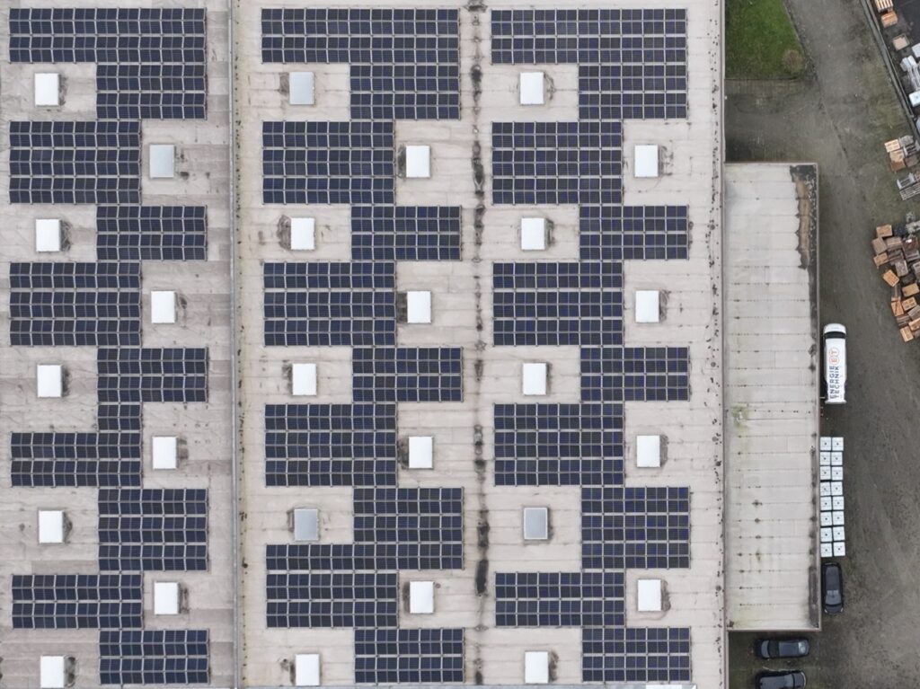 Solarmodule bedecken Fabrikdach, rechteckig angeordnet, mit grauem Gebäude daneben und parkenden Autos.
