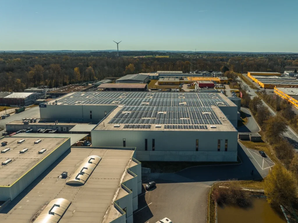 Dach des Industriegebäudes, bedeckt mit Solarpaneelen, in landschaftlicher Umgebung mit Windturbine.