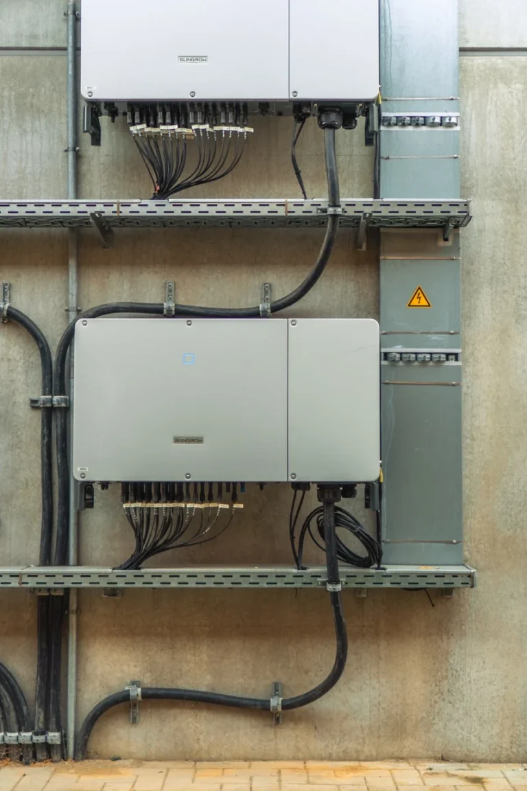 Solaranlage bedeckt Dach eines großen Gebäudes, umgeben von industrieller Landschaft und Wald. Gehäuse hängen, organisiert Kabel, an einer grauen Betonwand. Warndreieck mit Blitzsymbol sichtbar.