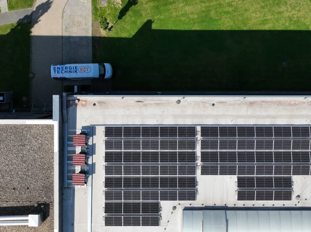 Solarzellen bedecken ein Flachdach, daneben steht ein Elektrotechnik-Firmenfahrzeug, umgeben von grünem Rasen.