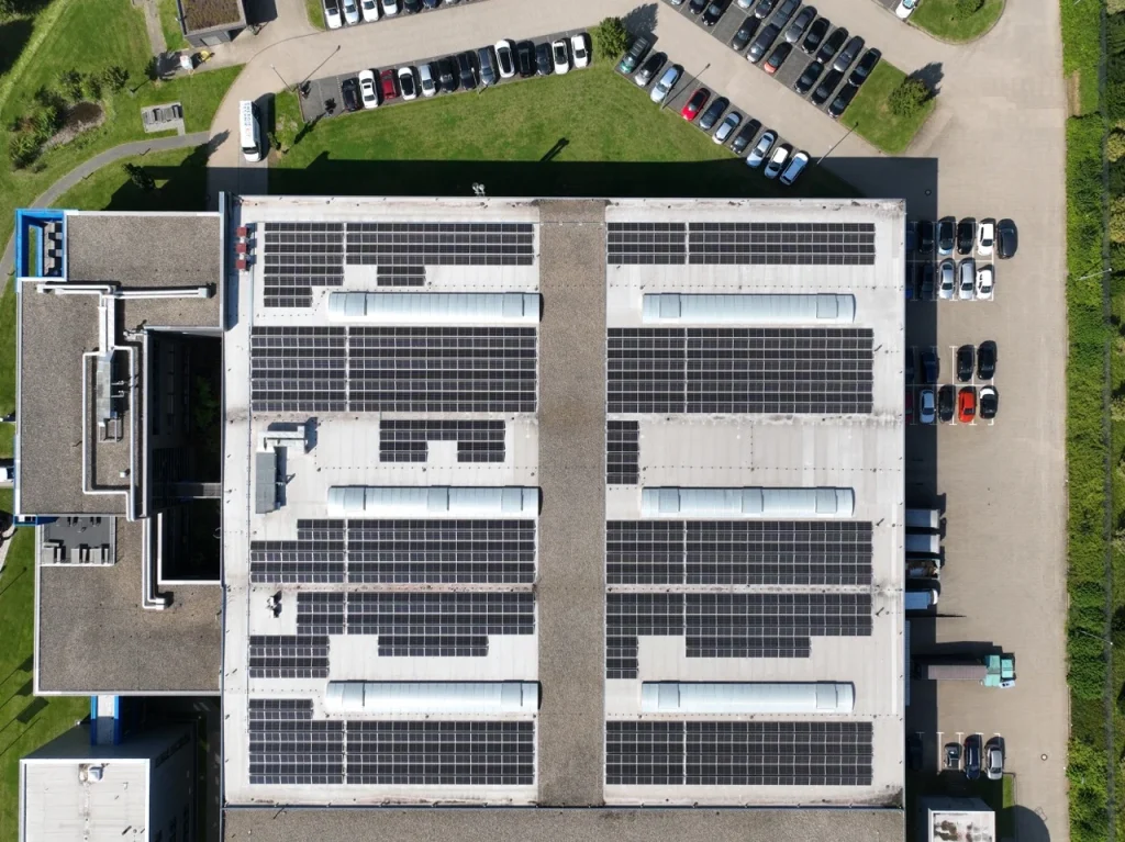 Solaranlagen bedecken Fabrikdach, Autos parken daneben, Umgebung mit grünen Rasenflächen.