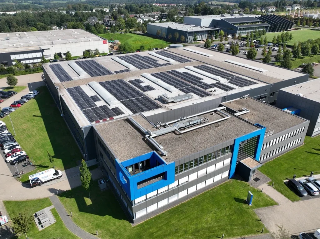 Großes Gebäude mit Solarmodulen auf dem Dach, umgeben von Grünanlagen und Parkplätzen.
