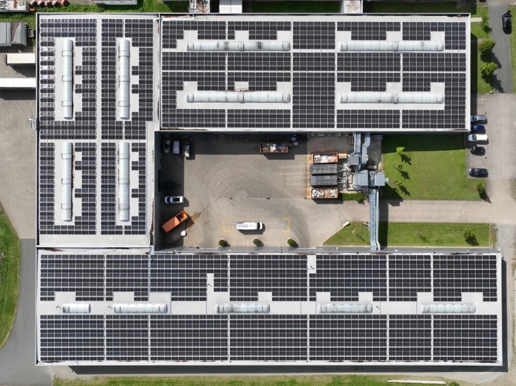 Solarpaneele bedecken das Gebäudedach, umgeben von Fahrzeugen und Grünflächen.