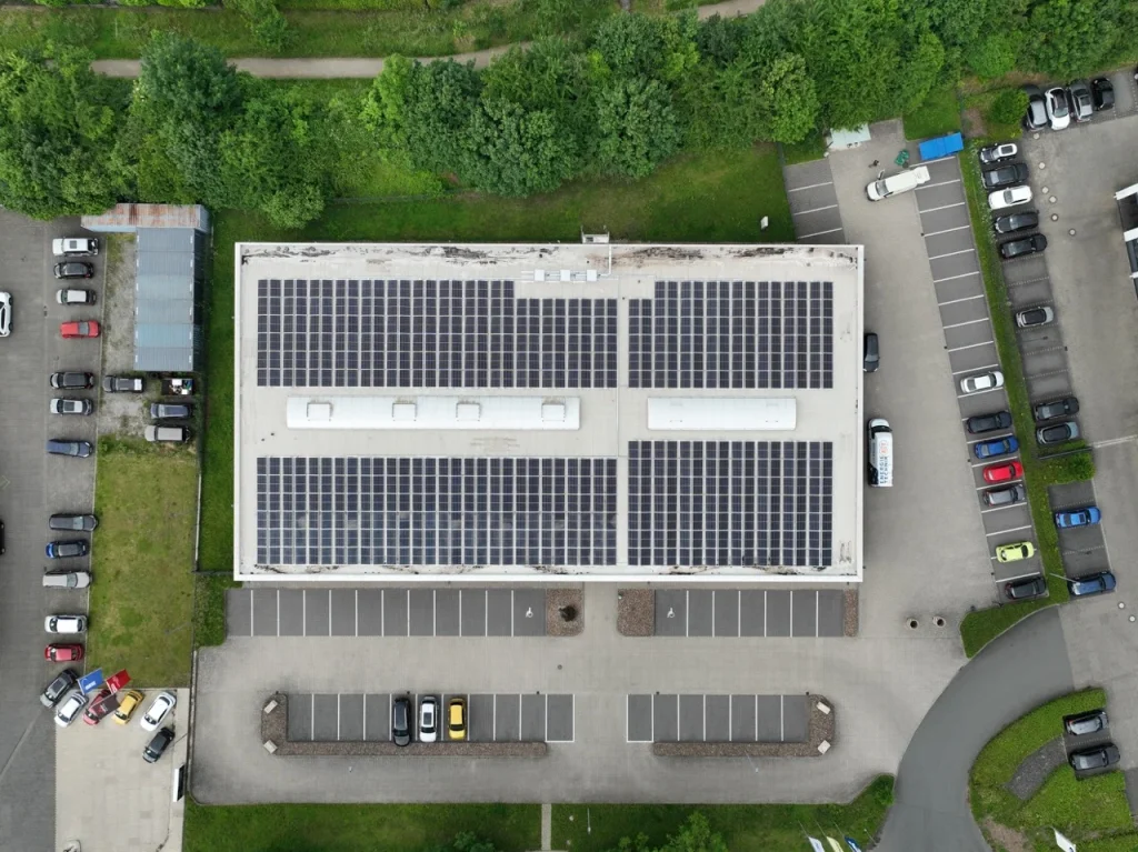 Gebäude mit Solarpanels auf dem Dach, umgeben von parkenden Autos und grüner Vegetation.