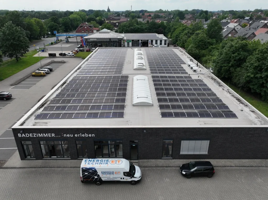 Solarmodule bedecken Dach eines Gebäudes, umgeben von Parkplätzen und Bäumen; Lieferwagen steht davor.