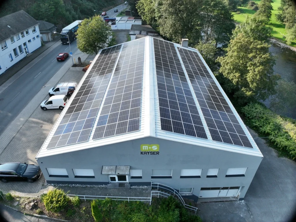 Gebäude mit Solarpanelen auf dem Dach, umgeben von Bäumen und parkenden Autos.