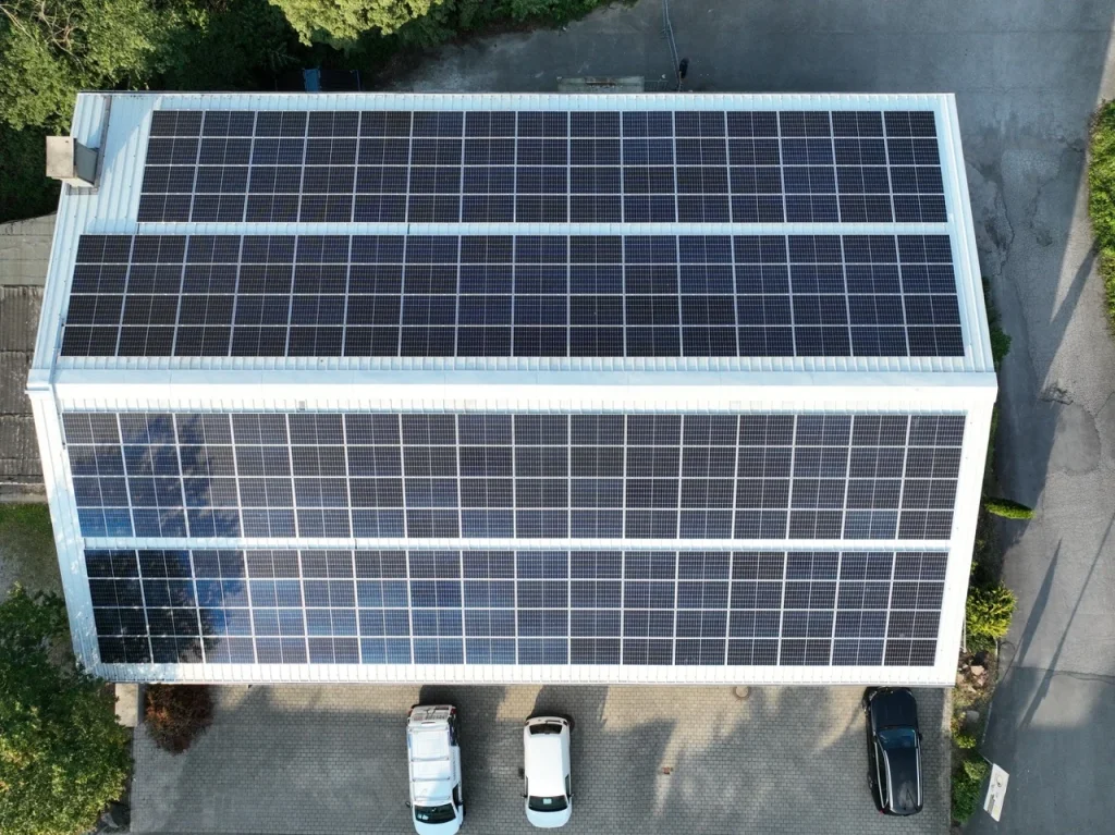 Solarpaneele bedecken Dach eines großen Gebäudes, umgeben von Bäumen, parkende Autos vorne.