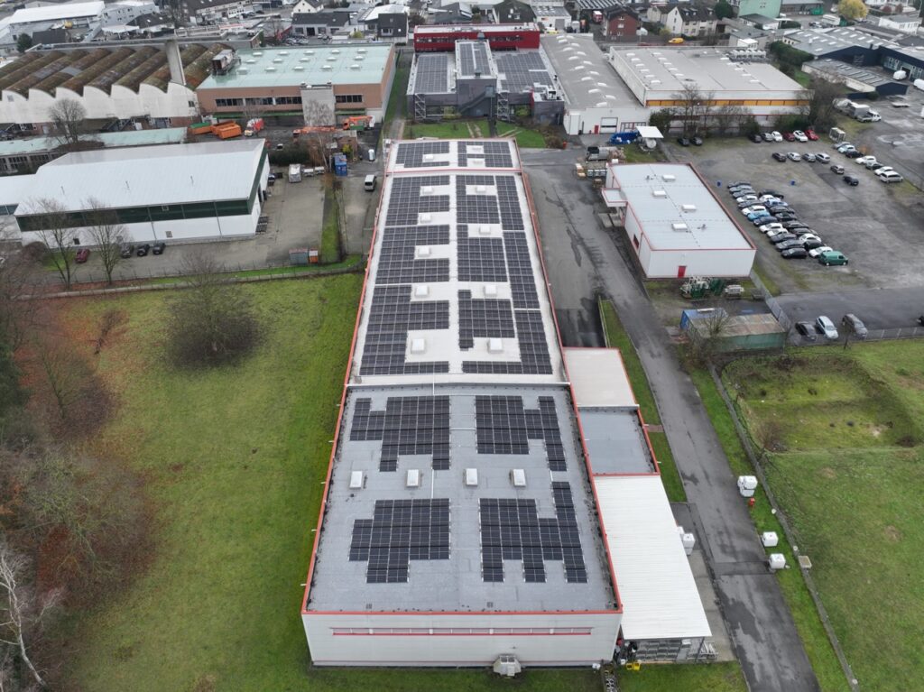 Dach zeigt große Solarpanel-Installation, umgeben von industriellen Gebäuden und Parkplätzen in grüner Umgebung.