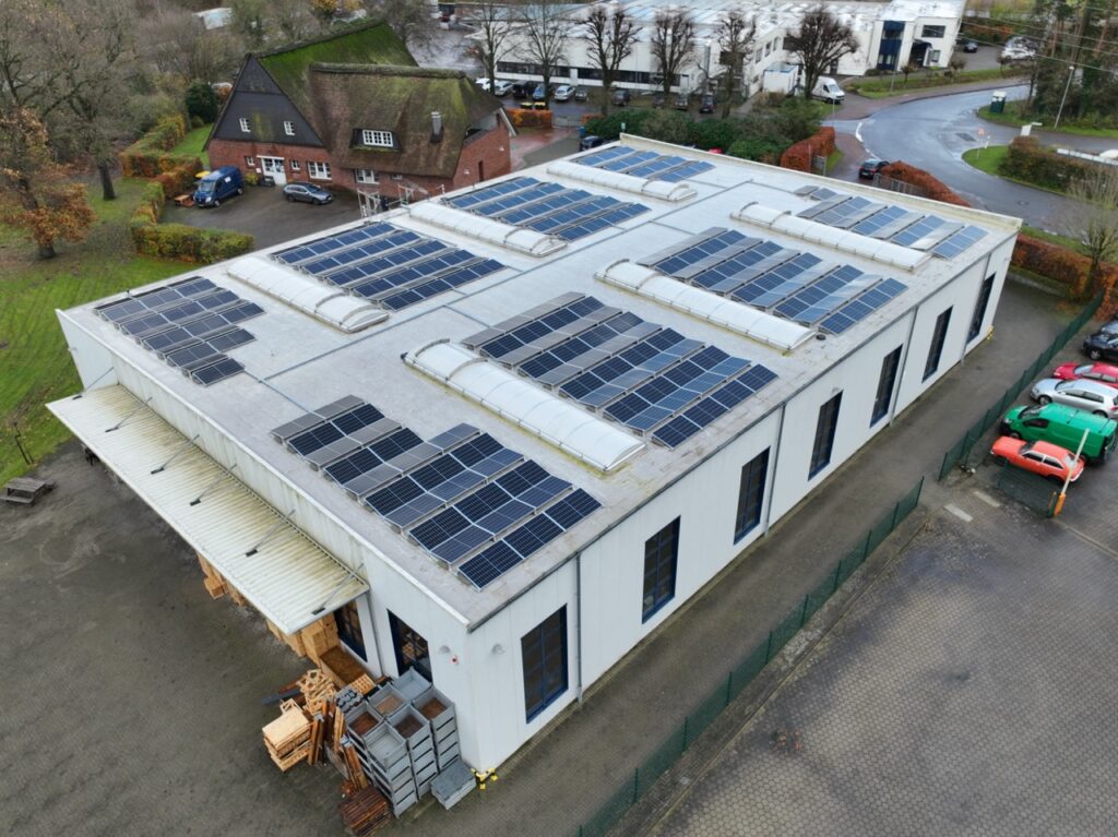 Flachdach mit Solarpaneelen, umgeben von parkenden Autos auf Asphalt und grünem Gelände.