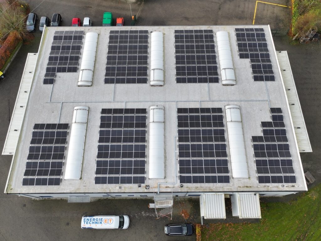 Flachdach mit Solarpaneelen, umgeben von parkenden Autos auf Asphalt und grünem Gelände.
