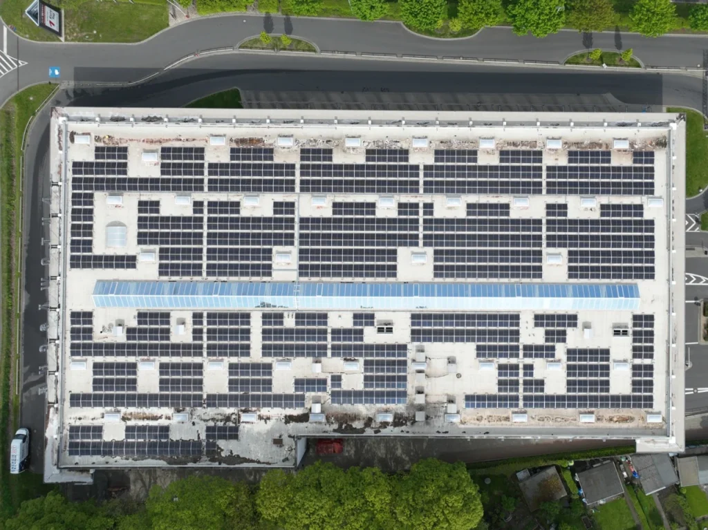 Flachdach mit Solarpaneelen, umgeben von parkenden Autos auf Asphalt und grünem Gelände.