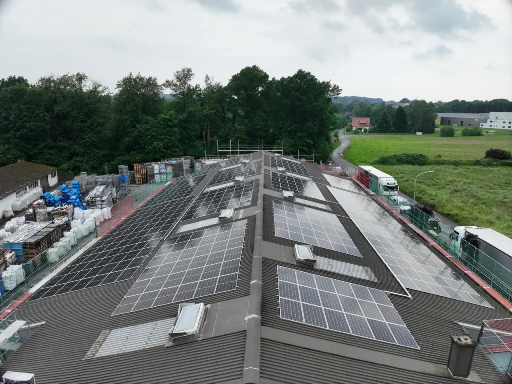 Solarmodule bedecken Hallendach in ländlicher Umgebung, umgeben von Bäumen, Lkw und Lagerflächen.