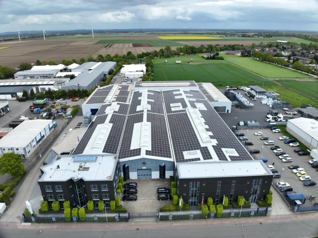 Großes Gebäude mit Solarpanels steht in einem Industriegebiet, umgeben von Feldern und Fahrzeugen.