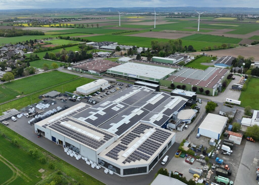 Industriebauten mit Solarpaneelen liegen in ländlicher Umgebung, umgeben von Feldern und Windkraftanlagen.