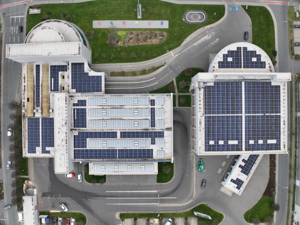 Gebäude mit Solarmodulen, aus der Vogelperspektive, umgeben von Straßen und Grünflächen.