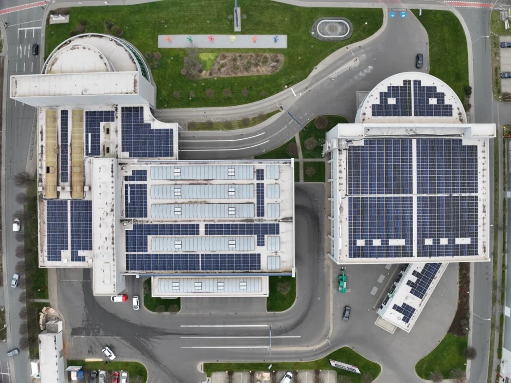 Gebäude mit Solarpanelen steht auf einem Parkplatz, umgeben von Straßen und grünen Flächen.