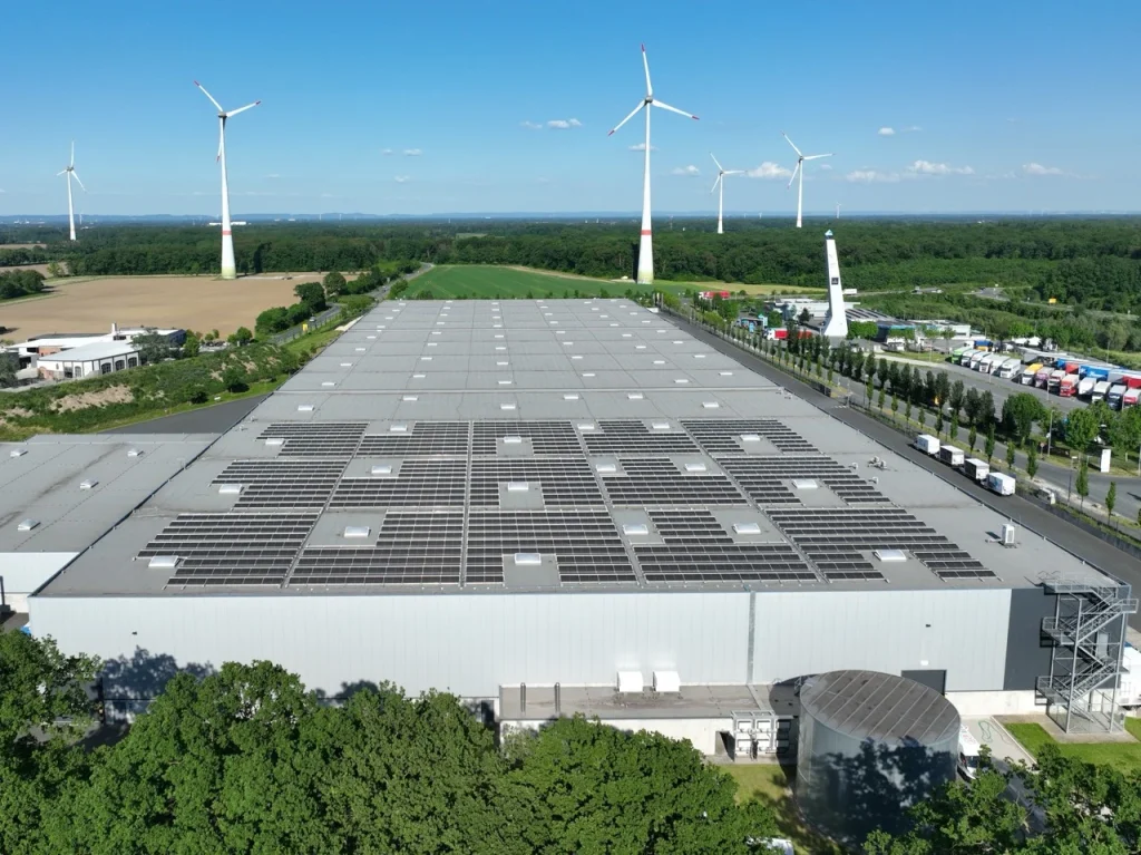 Dach eines Industriegebäudes trägt Solarpanels, umgeben von Windrädern und Grünflächen.