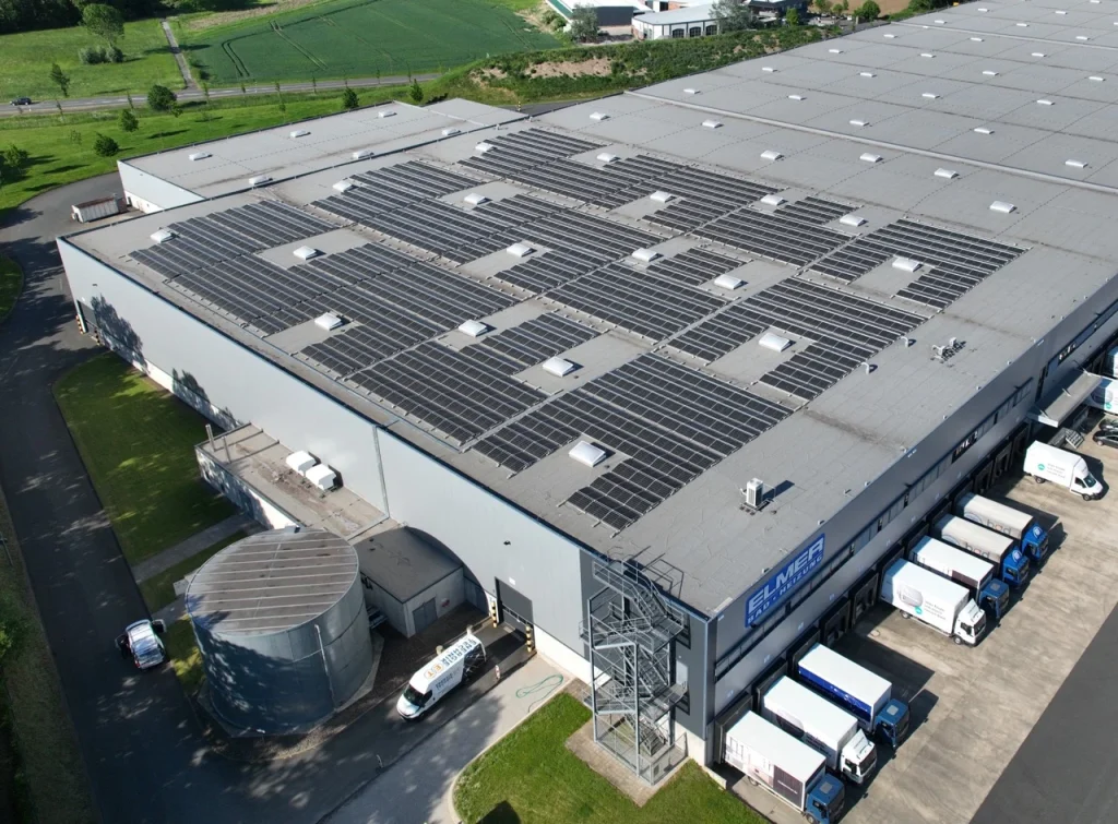 Das Gebäude trägt Solarpaneele und befindet sich neben parkenden Lastwagen in industriellem Umfeld.