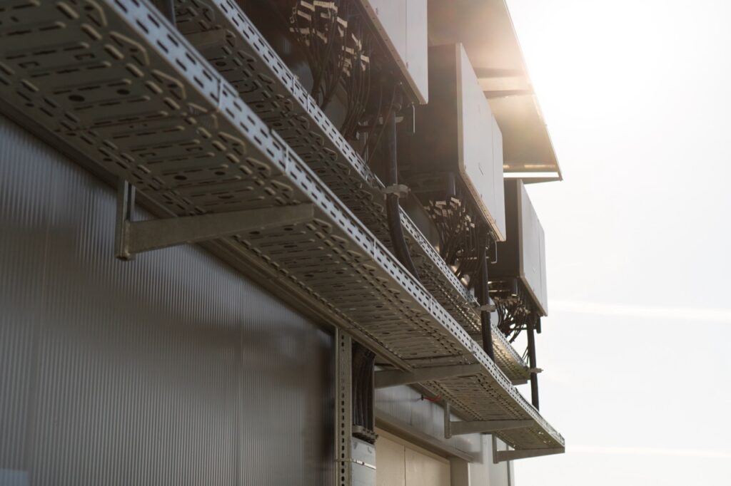 Metallplattform trägt elektronische Geräte; Kabel hängen herunter; im Sonnenlicht außen an Gebäude montiert.