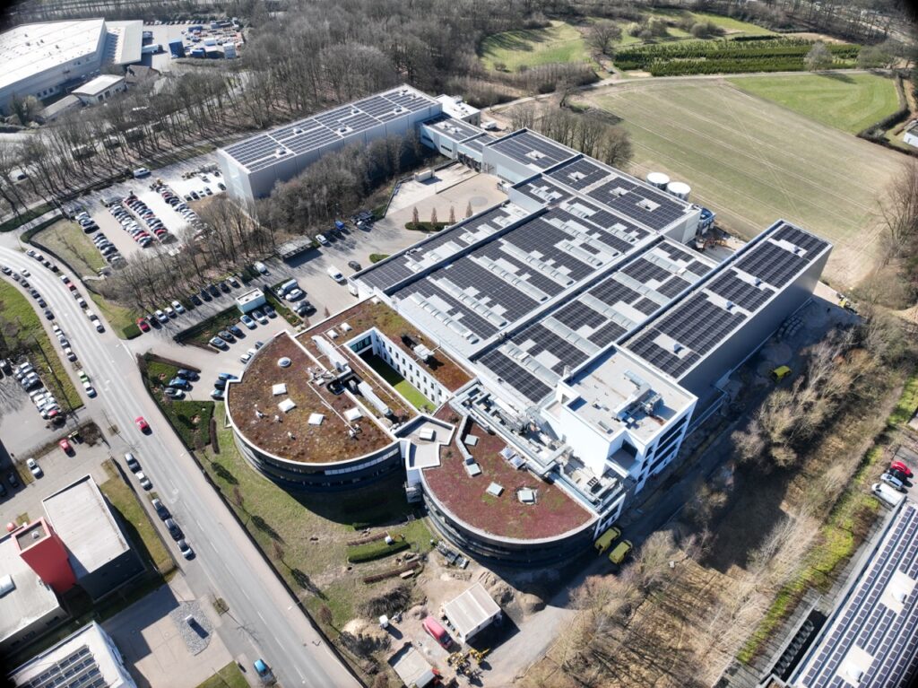 Großes Gebäude mit Solarpanels, umgeben von Parkplätzen und Feldern, aus der Luftansicht.