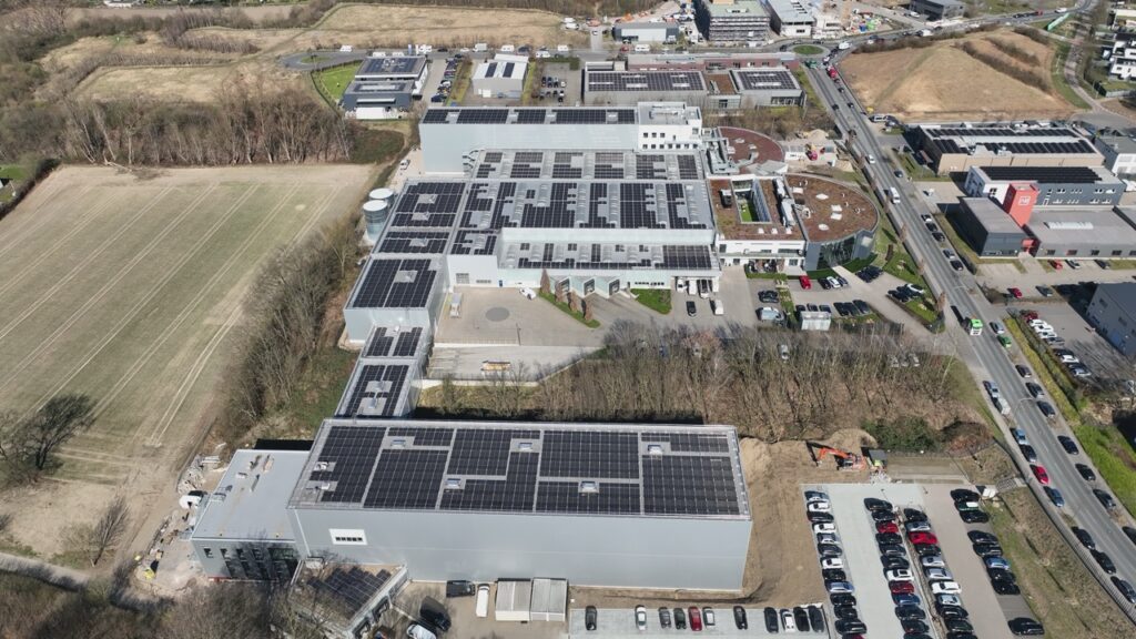 Industriegebäude mit Solarpanelen auf dem Dach, umgeben von Straßen und bewaldeten Flächen.