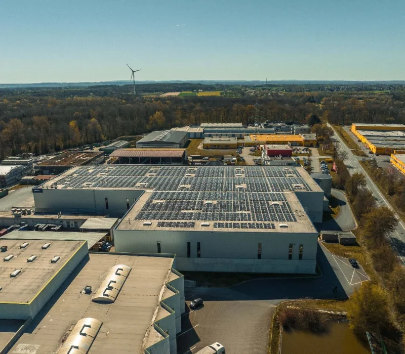 Dach des Industriegebäudes, bedeckt mit Solarpaneelen, in landschaftlicher Umgebung mit Windturbine.